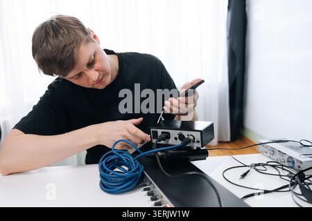 Fokussierter Mann richtet Audiogeräte ein und verbindet Kabel mit der Soundschnittstelle des Smartphones. Helles, natürliches Licht in einem modernen Studio. Technische c Stockfoto