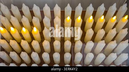 Viele elektronische weiße LED-Wachskerzen ohne Flammen stehen in einer Reihe und leuchten jeweils in warmgelbem Licht. Die Anordnung bildet ein einfaches und einheitliches Muster Stockfoto