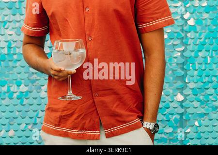Die Person hält ein Glas Wasser, während sie vor einer blauen dekorativen Wand bei einem gesellschaftlichen Treffen steht. Mann, der ein Glas mit Wasser hält. Einstellung sh Stockfoto