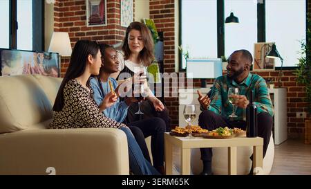 Freunde lachen, trinken Rot- und Weißwein, reden über ihr Leben im gemütlichen Wohnraum, Kollegen verbringen Zeit miteinander, essen von Wurstwaren zu Hause, zoomen in der Aufnahme. Stockfoto