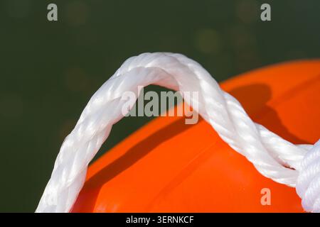 Weiße Seilschlaufen um die leuchtend orange Boje. Bootssicherheitsausrüstung, farbenfroh. Starkes, verdrehtes Kabel vor dunklem Wasser. Nahaufnahme hochgradig Stockfoto