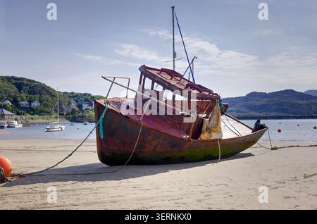 Dieses Küstenfoto zeigt ein verrostetes, stillgelegtes Fischerboot, das bei Ebbe an einem Sandstrand stark geneigt ist. Stockfoto
