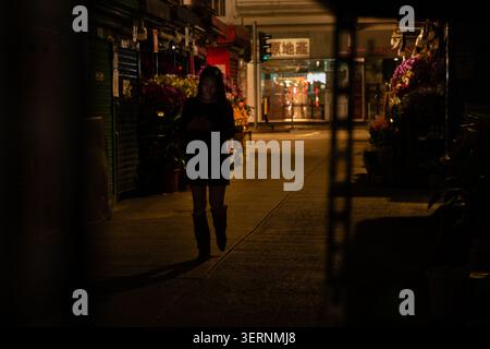 In Wan Chai, Hong Kong Island, läuft eine Frau nachts entlang einer schwach beleuchteten Straße, die von Blumenständen gesäumt ist, und hält ein Smartphone in chinesischer Sprache Stockfoto