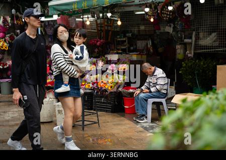 Fußgänger passieren den Jia Jia Blumenstand am Blumenmarkt in Prince Edward, Hongkong, während ein älterer männlicher Verkäufer neben bunten Blumensträußen sitzt Stockfoto