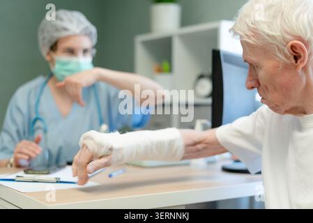 Senior-Mann, der sich während der ärztlichen Beratung an den Arzt wendet. Älterer Patient mit bandagierter Hand, der in der Klinik professionelle Betreuung sucht. Knochenfragilität und Osteoporose sorgen für ältere Menschen Stockfoto
