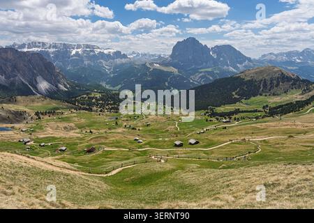 Blick aus einem hohen Winkel auf gewundene Wege und kleine Holzhütten, die über ein ausgedehntes grünes Alpental unter einem dramatischen Himmel mit felsigen Berggipfeln verstreut sind, Stockfoto