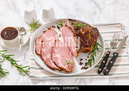 Kasseler Nacken, Kasseler Kamm Gourmet-Gericht mit zartem, geschnittenem Schweinehals auf einem weißen Teller mit frischem Rosmarin und köstlicher Sauce auf Marmortisch Stockfoto