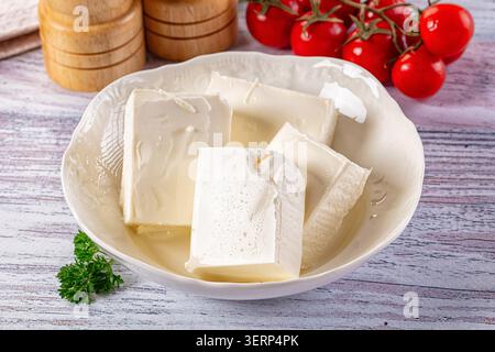 Frische weiße Feta-Käseblöcke in einer weißen Keramikschale. Hellrote Kirschtomaten auf Weinrebe, grüner Petersilienzweig sorgen für einen lebendigen Farbkontrast. Stockfoto