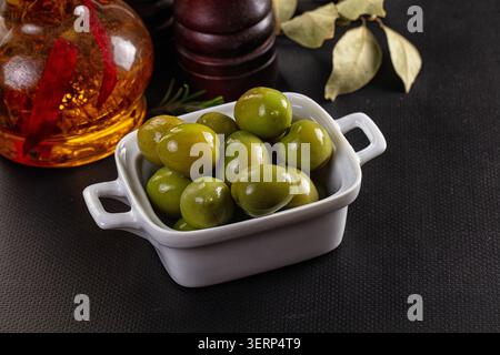 Frische grüne Oliven in einer kleinen weißen Keramikschale, begleitet von einer Flasche Chili-Olivenöl Stockfoto