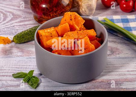 Gebackene, orangene Kürbisstücke, gewürzt mit Gewürzen in einer grauen Keramikschale. Gesundes vegetarisches Gericht auf hellem Holzhintergrund mit frischem Gemüse wie Bas Stockfoto