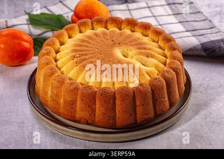 Geschäumter Kuchen mit geriffelten Kanten auf Keramikplatte, begleitet von frischen Orangen-Aprikosen und grünen Blättern auf kariertem Tuch, weiches natürliches Licht Stockfoto