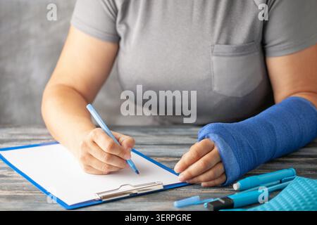 Frau mit blauem Abdruck auf dem Arm schreibt Versicherung mit einem Stift auf Papier auf grauem Hintergrund. Das Mädchen trägt eine gebrochene Hand Stockfoto