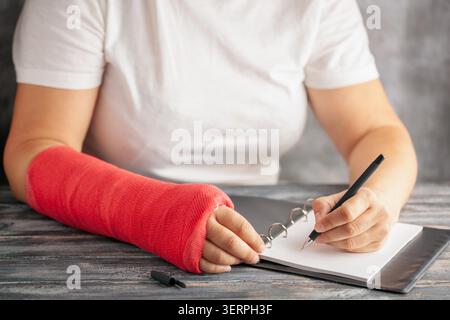Frau mit einem Gips am Arm schreibt Versicherung mit einem Stift auf Papier auf grauem Hintergrund. Das Mädchen trägt eine gebrochene Hand Stockfoto