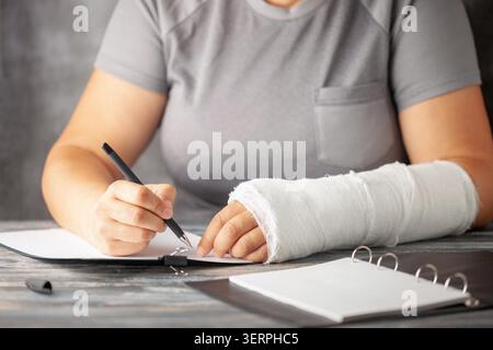 Frau mit einem Gips am Arm schreibt Versicherung mit einem Stift auf Papier auf grauem Hintergrund. Das Mädchen trägt eine gebrochene Hand Stockfoto