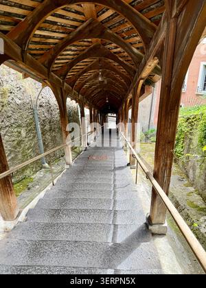 Eine historische überdachte Holztreppe mit bogenförmigen Holzbalken und abgenutzten Steintreppen führt durch eine tunnelartige Passage, flankiert von einem verwitterten Steinwal Stockfoto