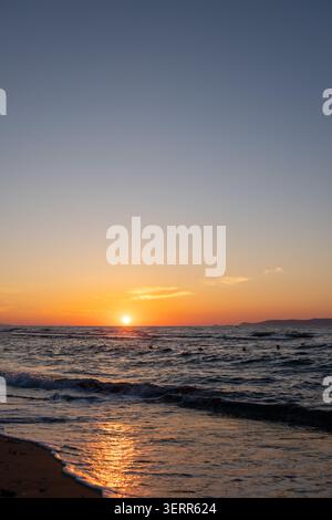 Eine vertikale Orientierungslandschaft, in der die Sonne den Horizont des Ozeans während eines goldenen Sonnenuntergangs berührt. Leuchtendes Orange und Gelb spiegeln schöne Farben wider Stockfoto