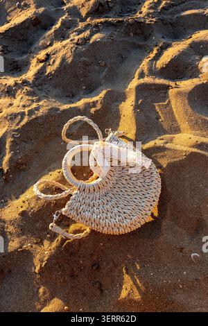 Ein Foto mit vertikaler Ausrichtung von einer stilvollen gestrickten Strohgeldbörse mit weißem Spitzenband auf einem Sandstrand. Die niedrige goldene Stunde Sonnenlicht krank Stockfoto