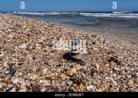 Eine Nahaufnahme einer kleinen Steinpyramide, die perfekt auf einem mehrfarbigen Kieselstrand balanciert ist. Im Hintergrund sanfte Meereswellen mit weißem Schaum Stockfoto