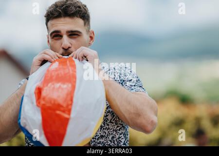 Junger Mann, der an einem sonnigen Sommertag draußen einen bunten Strandball aufbläst Stockfoto