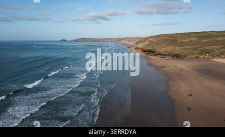 Blick aus der Vogelperspektive auf den langen Sandstrand und die Wellen mit Menschen, die entlang der Küste und der grasbewachsenen Klippen laufen, Perranporth, Cornwall, Großbritannien Stockfoto