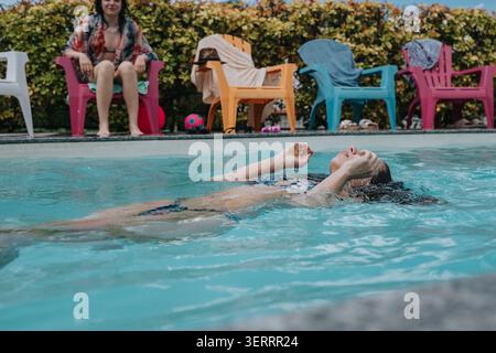 Frau schwimmt auf dem Rücken in einem Garten-Swimmingpool mit Freunden, die zuschauen Stockfoto