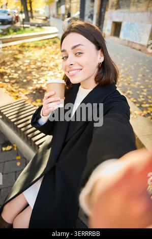 Junge Frau, die Selfie mit Kaffee zum Mitnehmen auf der sonnigen Herbststraße macht, Porträt, in schwarzem Mantel in die Kamera lächelt. Ideal für den urbanen Lebensstil, soziale Medien, Café-Werbung und Werbung Stockfoto