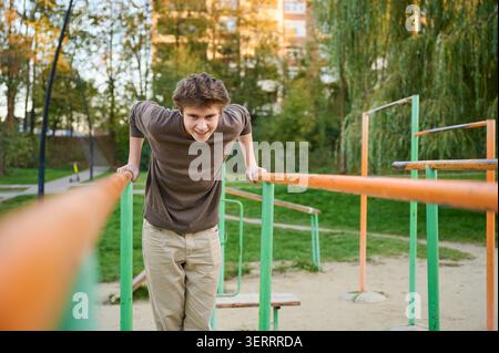 Weißer athletischer Mann, der parallele Dips mit kontrollierter Haltung durchführt, Calisthenics-Park-Umgebung im Freien mit orangegrüner Ausrüstung, intensivem Blick, Wohnblöcken und Bäumen im Hintergrund, Stockfoto