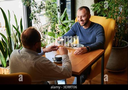 Zwei Männer unterhalten sich bei Kaffee am Kaffeetisch, freundliche Besprechung oder Geschäftsgespräche, entspannte Gespräche in modernem Innenraum mit Pflanzen und Tageslicht Stockfoto