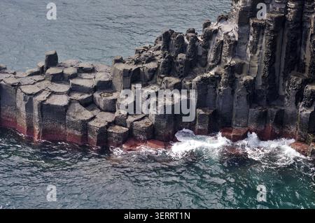 Nahaufnahme der Basalt-Säulenverbindungen, die durch vulkanische Aktivität auf der Insel Jeju in Südkorea entstanden sind, mit Wellen, die gegen die zerklüfteten Küstenfelsen schweben. Stockfoto