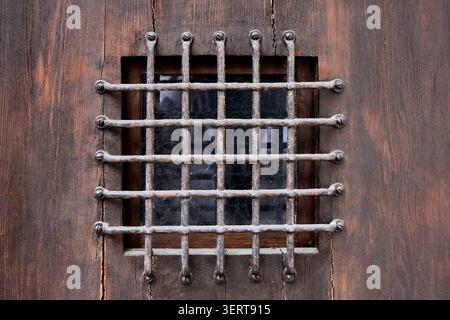 Kleines quadratisches Fenster mit einem schützenden schmiedeeisernen Grill an einer alten verwitterten braunen Holztür. Stockfoto