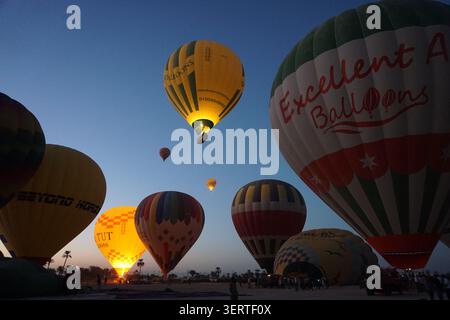 Heißluftballons bereiten sich auf den Start bei Sonnenaufgang mit Palmen und Crew in Luxor vor (sichtbare Firmennamen) Stockfoto