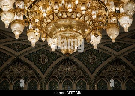 Blick auf den massiven, reich verzierten Kristalllüster, der von der aufwändig gemusterten Decke in der Sultan-Qaboos-Moschee in Muscat, Muscat G, hängt Stockfoto