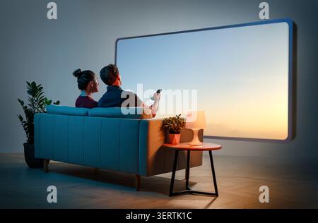 Ein junges Paar genießt einen entspannenden Abend zusammen auf einem stilvollen blauen Sofa in einem modernen Wohnzimmer und schaut einen großen, an der Wand montierten Fernseher oder einen Fernseher Stockfoto