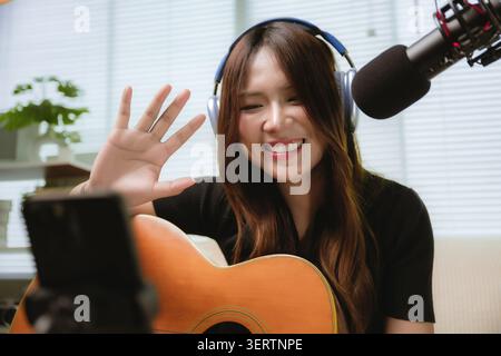 Asiatische junge Frau lächelt, während sie winkt, Kopfhörer und Gitarre trägt. Sie streamt Live-Musik in einem Heimstudio mit Smartphone-Aufnahme. Studio-Licht Stockfoto