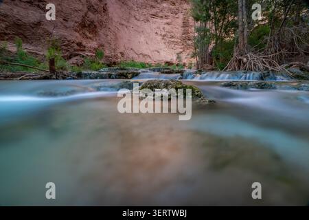 Blick auf das türkisfarbene Wasser des Havasu Creek in der Nähe der Havasu Falls, die über moosige Felsen mit roten Schluchtwänden und üppigen Bäumen in Supai, Arizona, United fließt Stockfoto