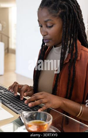 Erwachsene afrikanische Frau, die im Heimbüro auf einer externen Tastatur schreibt, mit Rostjacke und goldenen Armbändern Stockfoto