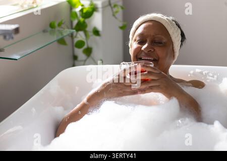 Ältere afroamerikanische Frau, die im Bad liegend mit Schaumglas mit bernsteinfarbener Flüssigkeit Stockfoto