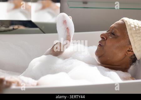 Ältere afroamerikanische Frau, die in der Wanne liegend, in Schaumtuch getränkt, Turban Spiegelregal, Chromhahn Stockfoto
