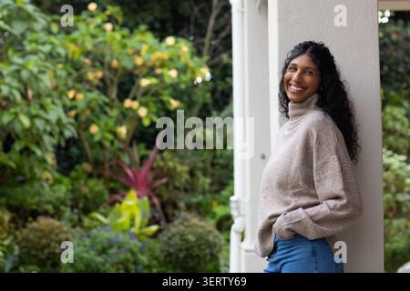 Afroamerikanerin lehnt sich an die weiße Säule, trägt beigefarbenen Pullover und Jeans, Kopierraum. Veranda, Garten, Outdoor, Denim, Freizeit, lockig, grüner Stockfoto