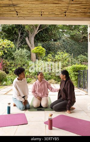 Verschiedene Freundinnen sitzen und plaudern auf überdachter Terrasse mit Yogamatten, Smartphone, Flaschen Stockfoto