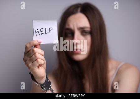 Menschenhandel. Frau mit Handschellen, die Papier mit Worthilfe auf grauem Hintergrund hält, selektiver Fokus Stockfoto