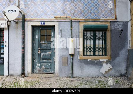 Nahaufnahme einer verwitterten Eingangstür und eines Gitterfensters unter dekorativen Azulejo-Fliesen an einer alten Wohnfassade in Lissabon, Portugal. Stockfoto