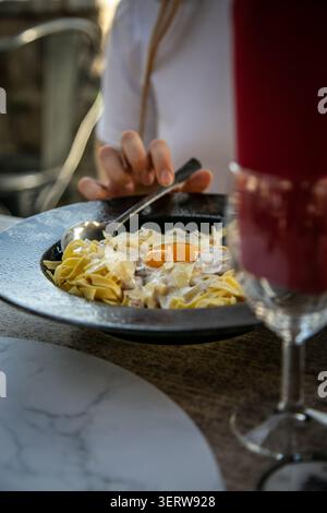 Eine Person isst Pasta Carbonara mit rohem Eigelb und geriebenem Parmesankäse, serviert in einer dunklen rustikalen Keramikschale in einem Restaurant Stockfoto