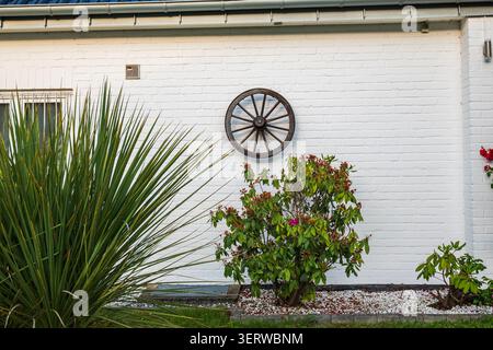 Hölzerne Wagenräder an der Wand eines weißen Backsteinhauses, rustikale Gartendekoration mit grünen Pflanzen und Ziersträuchern, Außendesign im Landhausstil Stockfoto