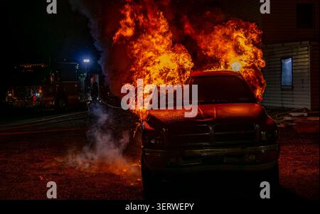 Flammen steigen aus einem Truck auf, der in der Nähe eines Hauses geparkt ist. Die Feuerwehrleute arbeiten daran, den Brand zu kontrollieren, während Rauch die Luft füllt. In den frühen Morgenstunden sind Rettungsfahrzeuge vor Ort. Stockfoto