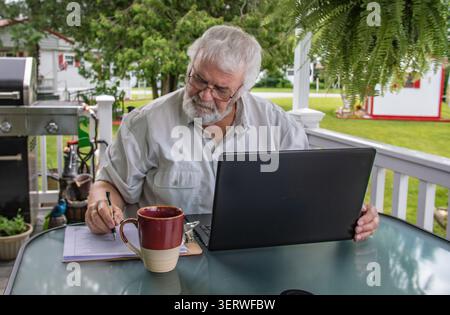 Ein Mann sitzt an einem Tisch auf einer Veranda im Hinterhof. Er sieht konzentriert aus, während er Notizen schreibt und einen Laptop benutzt. Ein Kaffeetasse sitzt neben ihm, während er bei Tageslicht arbeitet. Stockfoto