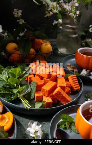 Orangen-Aprikosenmarmelade-Süßigkeiten mit frischer Minze auf einem dunklen Teller, neben warmem Tee in Kürbisform, die ein einladendes Frühlingsdessert schaffen Stockfoto