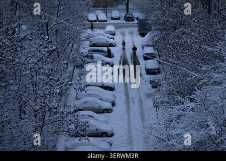 Eine ruhige Winterszene mit schneebedeckten Autos, die entlang einer engen, von Bäumen gesäumten Straße parken. Ein paar Leute laufen mitten auf der Straße, Leave Stockfoto