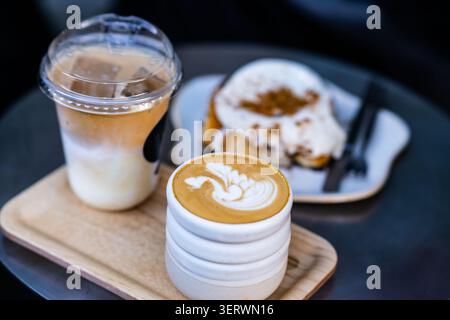 Eine gemütliche Kaffeepausen-Szene mit einem Latte mit komplizierten Schaumkunstwerken, einem Eiskaffee und einem Teller mit mit Zimt bestäubten Pfannkuchen. Stockfoto