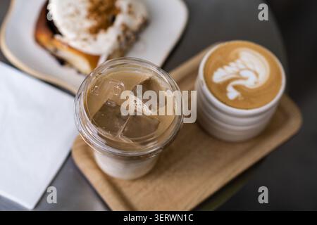 Eine gemütliche Café-Szene mit einem Latte mit komplizierten Schaumkunstwerken, einem Eiskaffee und einem überzogenen Dessert, alles auf einem Holztablett angeordnet. Stockfoto